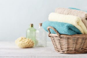 Four folded bath towels in a basket with a sponge and two glass bottles in the background.