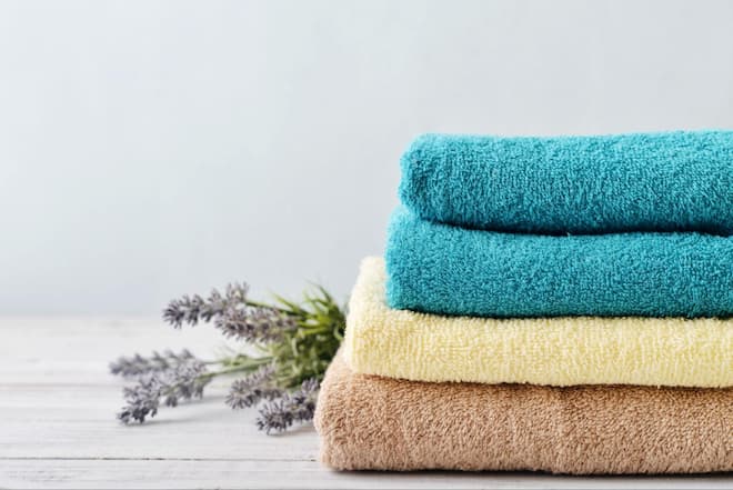A stack of folded towels, one brown, one pale yellow, and two turquoise. A sprig of fresh flowers sticks out from the bottom towel.