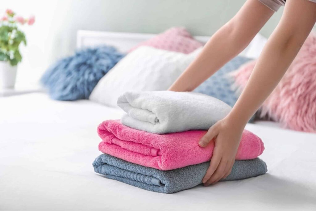 A woman places a stack of three plush towels on a bed.