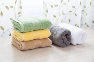 A pile of three folded towels, one brown, one yellow, and one green, stacked next to a rolled gray towel and a rolled white towel.