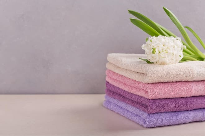 A stack of four towels with a white flower on top. The stack features an off-white towel on top of three pastel towels in pinks and purples.