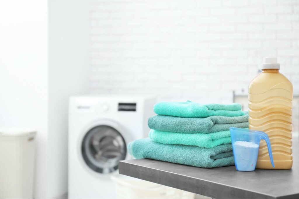 A stack of clean teal towels on a table with a cup of detergent and baking soda, a washing machine in the background.